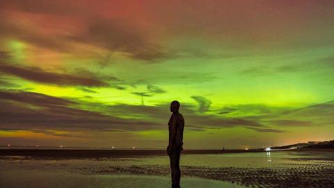 Vivid green and yellow lights are visible in the night sky above a beach, with an iron statue of a man visible in the foreground. 
