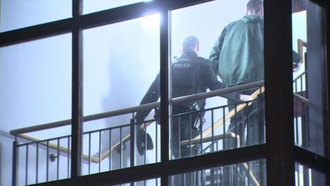 Two male police officers walk up the steps of the flat block. They are in green police uniforms and have their backs to the camera