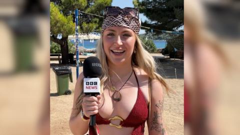 A girl with long blonde hair and light eyes stands in front of a beach. She is wearing sunglasses and a bandana on her head, a chunky brown necklace, and a red bikini top. She is holding a BBC News mic.