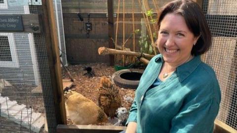 Clare Howard wears a blue shirt and by a chicken coop with her chickens in view