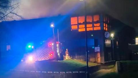 A fire engine and firefighters standing outside a large building that is on fire