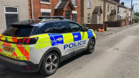 A police car is parked on the pavement outside a row of houses in Tibshelf