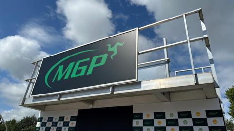 The Manx Grand Prix podium, which is a platform with railing around it and a large board featuring the green MGP logo on the front. The sky above is blue with white fluffy clouds.