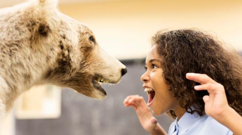 A girl with curly brown hair pretending to roar at a stuffed bear in a museum.