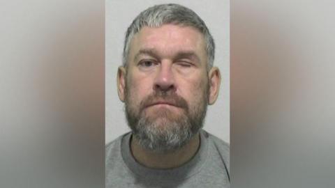 Mugshot of Wayne Kilty. He has a thick grey beard and short grey hair. His left eye is closed.