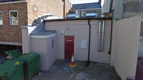 The rear of the Costa Coffee shop, which is a cream building with a red door with the word Costa written on it in white. There is a traffic cone in front of the door and large green bins to the left.