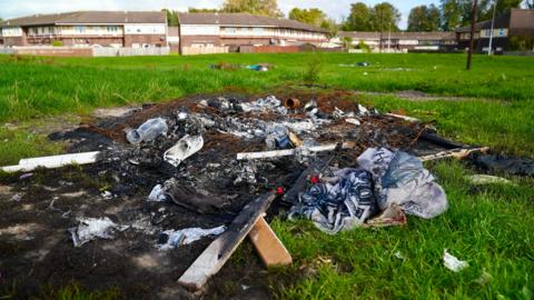 A pile of burnt rubbish in the foreground, including what's left of a burnt mattress, you can see more rubbish and then homes in the distance.