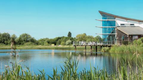 A lake with a white and glass building to the right. People are siting on some decking outside.