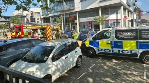 A fire engine and police van parked outside a post office. There is another white car and black car parked next to them. There are emergency workers standing outside.