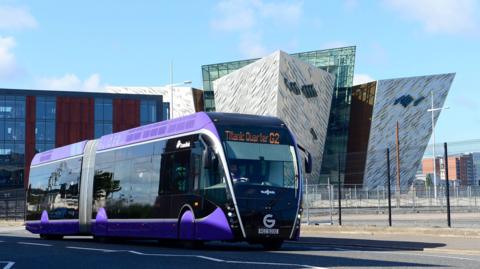 A glider is parked on a road. It's a large purple bus. Titanic Quarter is displayed on the digital board in front of the bus. A Titanic museum is behind the bus. The building has sharp edges at each side to look like a ship.