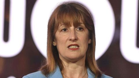 Chancellor Rachel Reeves, wearing a blue jacket, stands in front of a bright white sign, with the o in the sign surrounding her head.