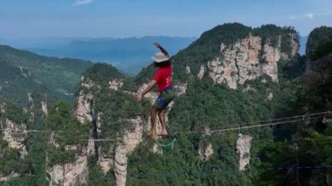 A man wearing a red t-shirt and hat balances on a slackline high above a gorge in China. In the background, tall rock faces covered in green trees reach far into the distance.