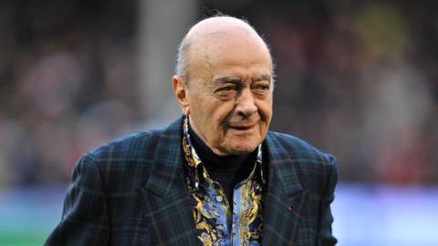Mohamed Al Fayed is slightly smiling, he is balding with grey hair and eyebrows and wearing a dark green tartan blazer and patterned yellow and blue shirt 