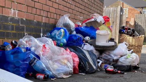 Bags of rubbish piled in a street against a red brick wall.