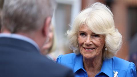 A picture of Queen Camilla taken over the shoulder of a man wearing a suit. She is wearing a blue dress.