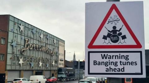 A red triangle warning sign that reads "Warning banging tunes ahead" with a cartoon man playing a drum set. The sign is in front of Glasgow's Barowland sign.