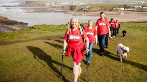 Several people wear matching red T-shirts with the words Heli Hike in white across them. They are walking up a green coastal hill. The tide is out behind them and there is a sandy beach surrounded by cliffs. They are all wearing trousers, some have coats tied around their waist, some have walking poles, and a couple of them have a dog on a lead.