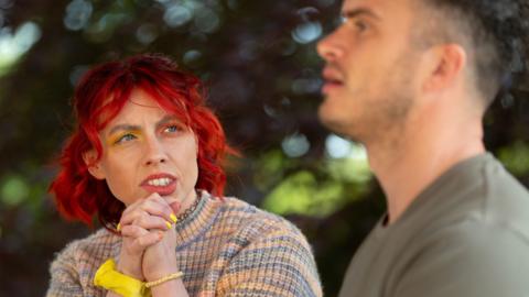Traitors contestant Jessie Stride is seen chatting to fellow faithful Matthew. Jessie has bright red hair, yellow and orange eyeshadow and is wearing a rainbow jumper. The background has trees.