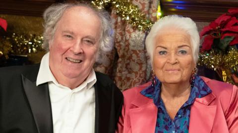 Nigel (Paul Bradley) with Pat (Pam St Clement) in character in the Queen Vic, in sitting and smiling in front of Christmas decorations