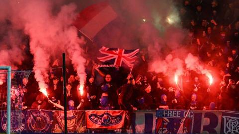 Football fans in a stand, covered with banners and with pyrotechnics and flares going off. Some fans are waving a Union Jack flag.