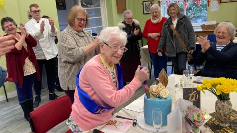 Tomasina Crowe is standing up, wearing a pink jumper and is cutting a blue birthday cake with a knife. She has friends and family behind her