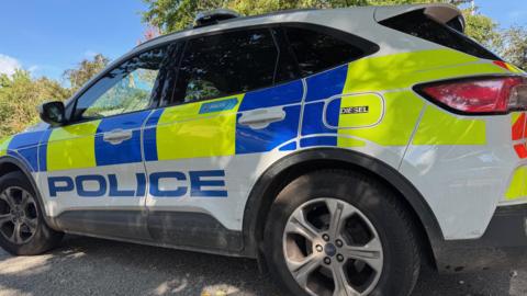 A stationary police car with gravel underneath it. There are trees behind it and it is a sunny day.