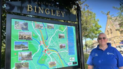 A middle-aged woman with short, cropped, grey hair. She wears a blue polo shirt. She is standing in front of a town sign for Bingley