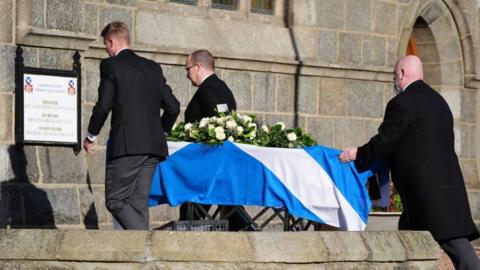 Three pallbearers take Alex Salmond's coffin into a church. The coffin is draped in a saltire and has flowers on top.