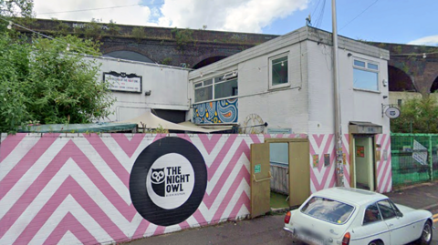 A view of the outside of the Night Owl. A wall at the front is painted white with pink chevrons. The rest of the building is white, with railway arches in the background