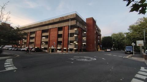 A car park with breeze block style walls - it is five levels high. A road and a roundabout is nearby with a sign with a blue 'P' for parking directing drivers to the car park.