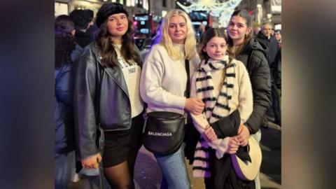 A woman in white jumper and jeans, three girls two in black coats, one in cream jumper