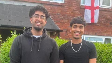 Two men stand outside a house smiling. An England flag is hanging out of one of the top windows of the house