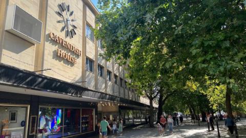 Cavendish House, a large department store building on Cheltenham's Promenade, glows in the sun. Shoppers are walking beneath leafy trees in the shade. There is a large clock on the front of the building.