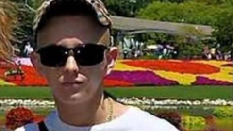 A teenager, wearing a plaid cap and dark sunglasses. He is also wearing a white t-shirt with black writing which says 'BOSS, HUGO BOSS' on it. A black bag strap in across his top. In the background are lots of brightly coloured flowers.