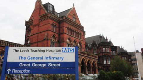The exterior of Leeds General Infirmary, as indicated by an entrance sign indicating the reception is ahead. The hospital building itself is a grand old Victorian structure.
