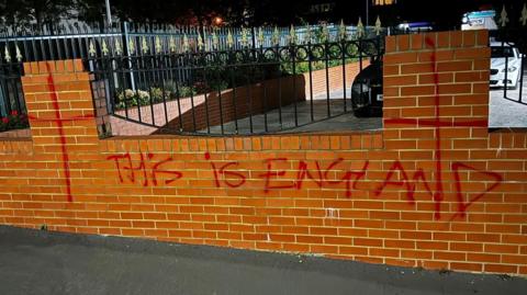 A brick wall that has "This is England" spray-painted in red on it. Behind the wall are two parked cars. The picture has been taken at night.
