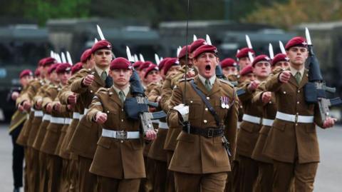 A group of uniformed soldiers march in tight formation across a paved surface. They wear brown military uniforms with red berets and carry rifles. The front-centre soldier leads with a commanding stance, mouth open as if shouting orders. Behind them, military vehicles are parked in a neat row.