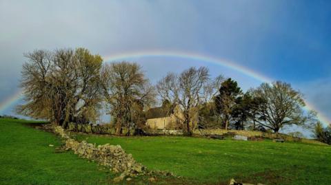 All Saints Church on top of a hill. It is surrounded by trees and fields. A rainbow fills the sky above the building.