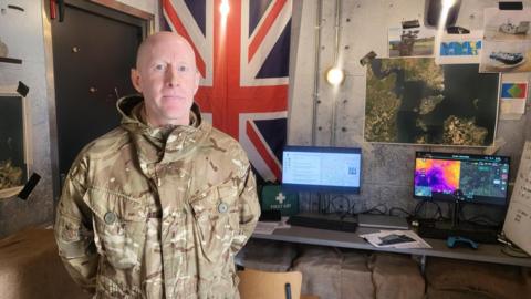 A white man in a green camouflage army outfit. He is standing in a bunker like room that is an immersive training exercise.  