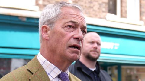Reform UK leader Nigel Farage wearing a suit and looking into the distance, in front of a building with a blue shop front in the background