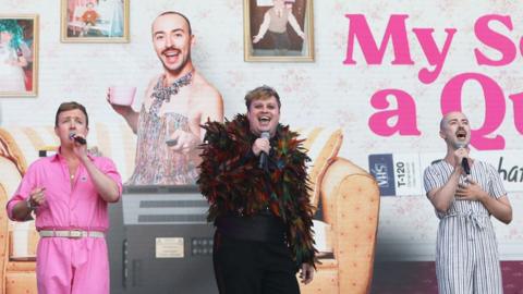 Three cast members from My Son's A Queer (But What Can You Do?) singing in front of a billboard with graphics from the show