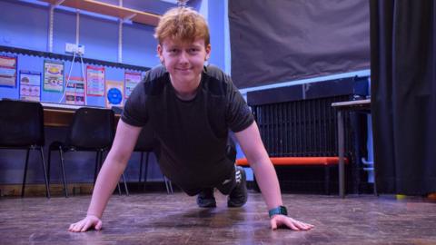 Adam is lying in the push-up position and is smiling at the camera, he is wearing a black t-shirt and black joggers with trainers. 