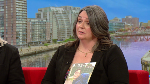 Louise is sitting on a red sofa in BBC Breakfast studios holding a photo of her son. She has a black jacket on and shoulder length brown hair.