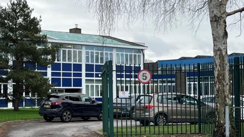 A school building with blue walls and large windows. A green metal fence surrounds the building.