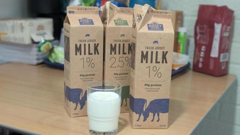 Three Jersey Dairy milk cartons. There is a glass of milk in front of it. Behind them are biscuits.