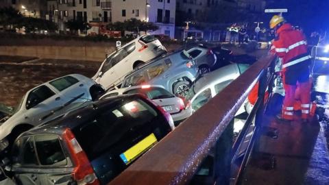  A fireman in a red suit with white reflective stripes and a yellow helmet looks down on a pile of cars, as water flows under them, at night-time