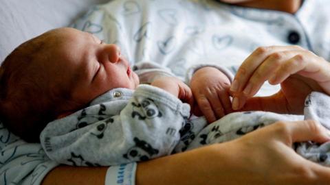 Generic image of a newborn baby resting in his mother's arms as she clutches his hand