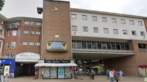 Broadgate House in Coventry city centre. It is a tall, brown brick structure with a clock on the outside and shops at the base. It is connected through a white/grey connecting passage to a neighbouring building.