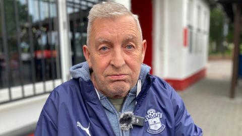 A man with short grey hair looks upset sat outside a red and white building. He is wearing a blue Southampton FC jacket and a grey hoodie.
