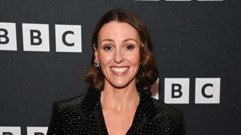 A woman in her fourties with brown hair in a curled and styles bob. She wears a black shirt or blazer with tiny rhinestones spread across it. Behind her is a black board with the BBC logo on it in white.